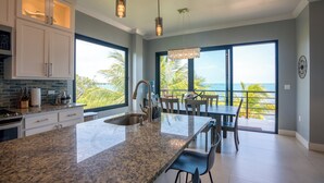 A modern kitchen with granite countertops, stainless steel appliances, and a large island. There is a dining area with a table and chairs, and a view of the ocean through large windows. - Sunset Gecko Condos - Unit 1A (Placencia)
