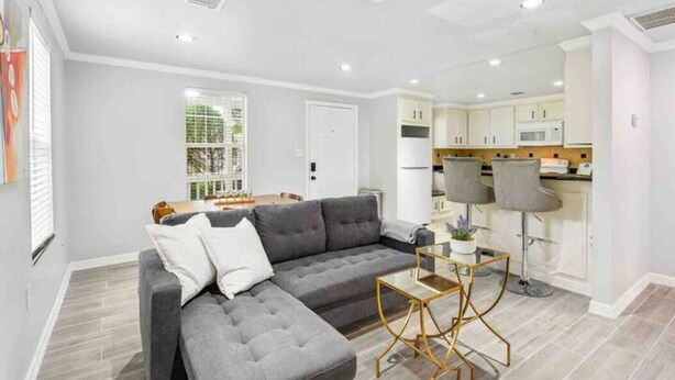 Una sala de estar moderna con un sofá seccional gris, una mesita de centro pequeña y una cocina con armarios blancos y un taburete de bar.