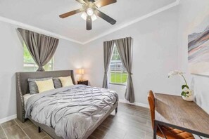 Une chambre à coucher comprenant un lit, un ventilateur de plafond, une table de chevet, une table avec une chaise et une fenêtre avec des rideaux.