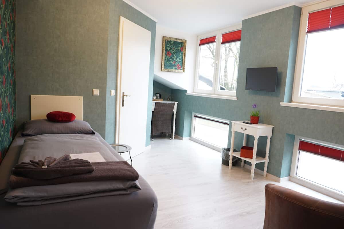 A bedroom with a bed, a small table, a chair, a TV, and a window with blinds.