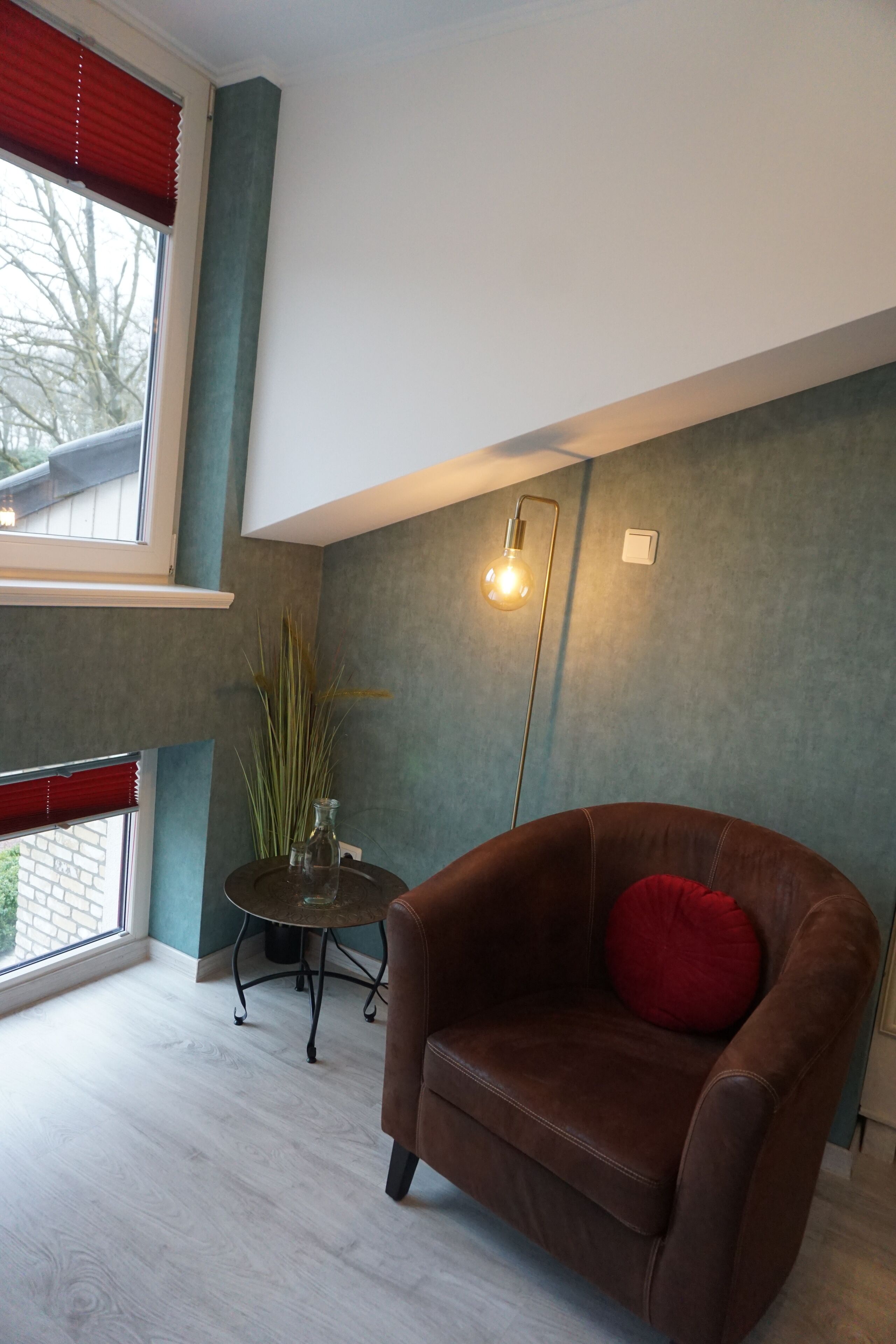 A room with a brown leather armchair, a small round table with a vase, a tall floor lamp, and a window with red blinds.