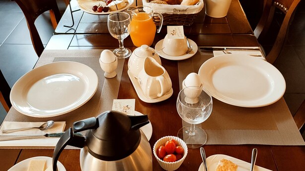 Una mesa de desayuno dispuesta con platos, cubiertos, una tetera, un cuenco de fruta y una cesta de pan.