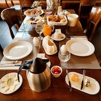 A breakfast table set with plates, cutlery, a teapot, a bowl of fruit, and a basket of bread.