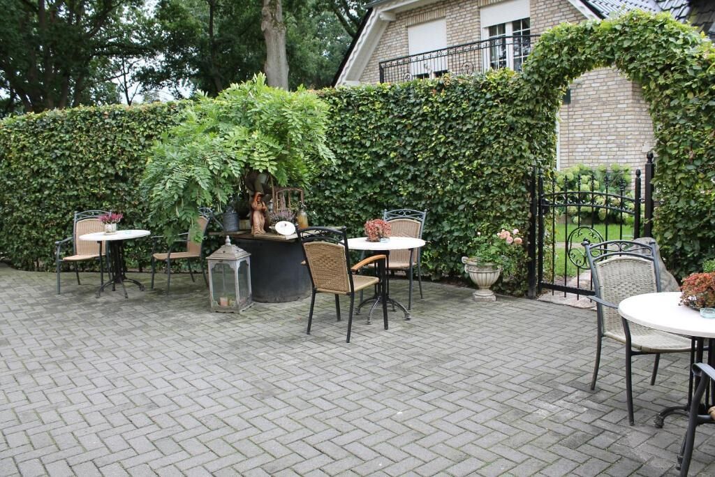 An outdoor patio with brick flooring, surrounded by lush greenery and a brick building.