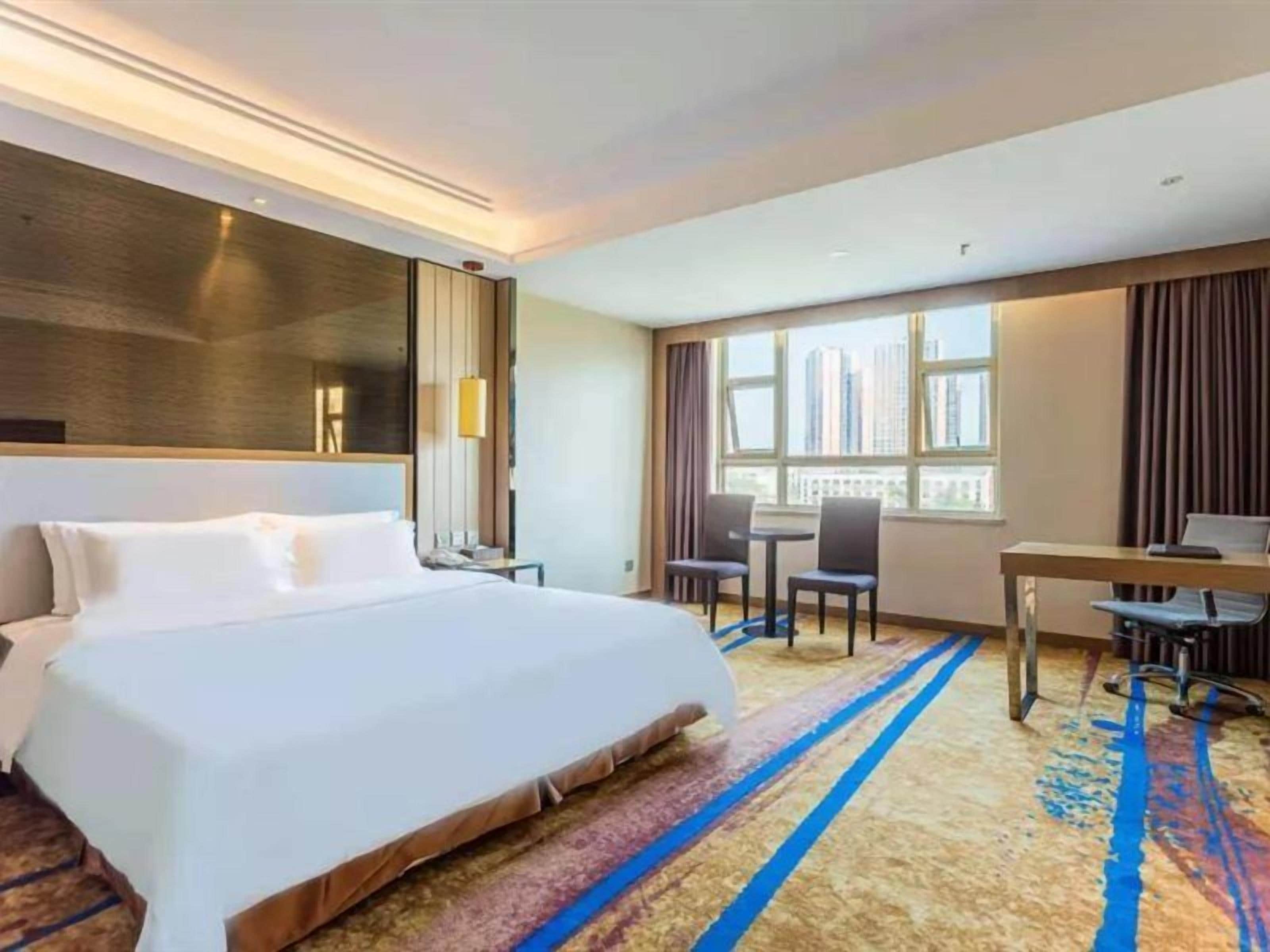 Quarto de hotel com uma cama grande, uma escrivaninha e uma cadeira. Há uma janela com vista para edifícios.