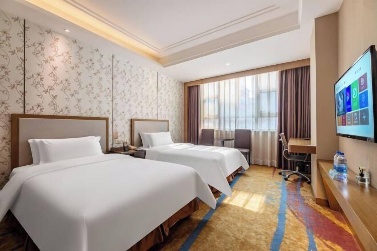 Ein Hotelzimmer mit zwei Betten, einem Schreibtisch, einem Stuhl, einem Fernseher und einem großen Fenster mit Vorhängen.