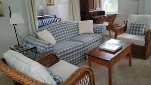 Een gezellige woonkamer met een blauw-witte geruite bank, een rieten fauteuil en een houten salontafel. Achterin staat een piano en er is een raam met gordijnen.