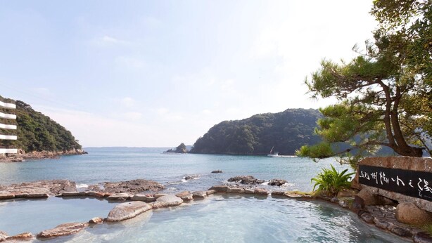 Una piscina cristalina con rocas en primer plano, un letrero con caracteres japoneses y vistas al mar y las colinas al fondo.