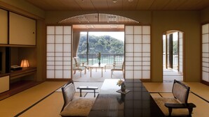 Una habitación japonesa tradicional con suelo de tatami, una mesa baja con sillas y vistas a una zona boscosa.