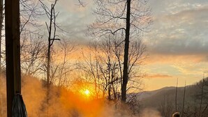 A hot tub in a natural setting with a sunset view, surrounded by trees and a wooden deck. - Whispering Falls Cabin - hot tub, views, 15 acres, minutes from Main St. (Saluda)