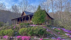 A wooden cabin with a porch, surrounded by a garden with purple flowers, set in a forested area. - Whispering Falls Cabin - hot tub, views, 15 acres, minutes from Main St. (Saluda)