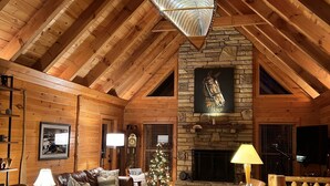 A cozy log cabin interior with a stone fireplace, leather furniture, and a wooden ceiling. - Whispering Falls Cabin - hot tub, views, 15 acres, minutes from Main St. (Saluda)