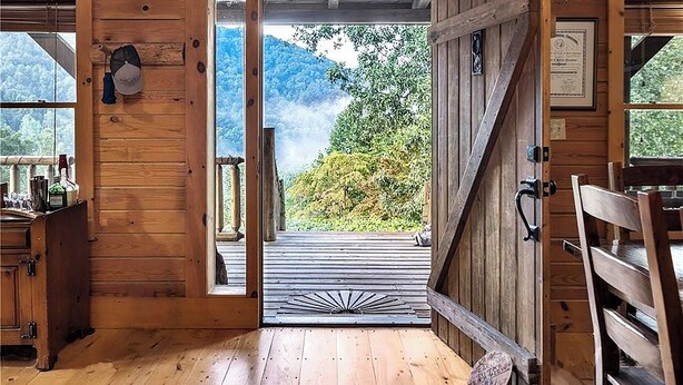 A wooden cabin interior with a large open door leading to a deck with a view of a forested mountain. - Whispering Falls Cabin - hot tub, views, 15 acres, minutes from Main St. (Saluda)