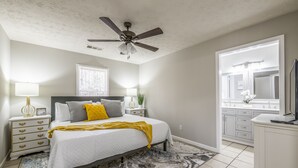 A bedroom with a bed, nightstands, a ceiling fan, a window, and a bathroom visible through an open door.