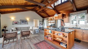 A spacious living area with a sofa, two wicker chairs, a coffee table, and a red rug. A kitchen with wooden cabinets and a large island is visible.