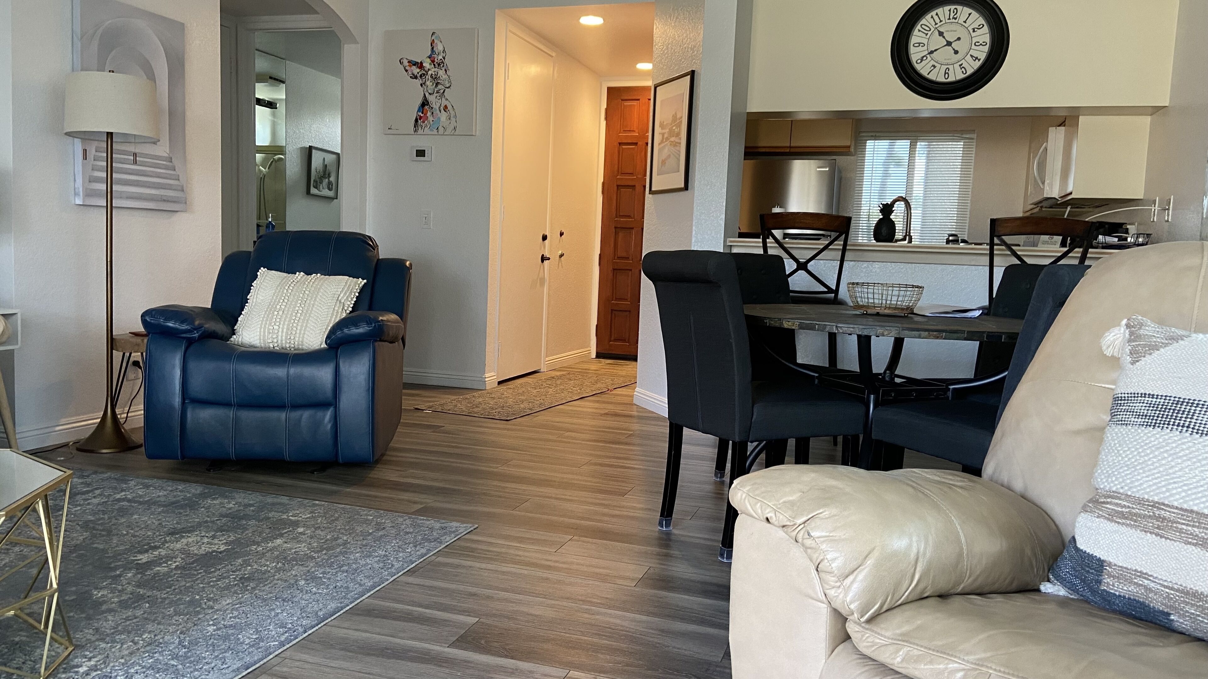 Sala de estar com uma poltrona de couro azul, um sofá bege e uma mesa de jantar com cadeiras. Uma área de cozinha ao fundo, com um relógio na parede.