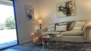 Ett vardagsrum med en beige soffa, ett litet runt soffbord och en lampa. Det finns en dörr som leder ut till ett utomhusområde.