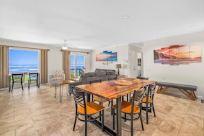 A spacious living area with a wooden dining table, comfortable seating, and a sofa. Large windows offer a view of the ocean.