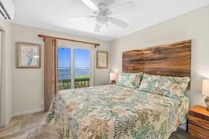 A bedroom with a large bed, a ceiling fan, a wooden headboard, a nightstand, and a view of the ocean through a sliding door.