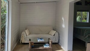 A cozy room with a wooden ceiling, a cushioned bench, a wicker table, and a large window. - The Surf Cottage - Beach and Jungle all in One!  An off the grid cottage. (Gregory Town)