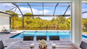 Zona de piscina exterior techada con vistas a un lago, terraza de madera y mobiliario exterior.