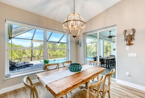 A dining area with a wooden table and chairs, a chandelier, and large windows overlooking a pool. - Waterfront Key West Style Home with Heated Pool Located in South Gulf Cove (Port Charlotte)