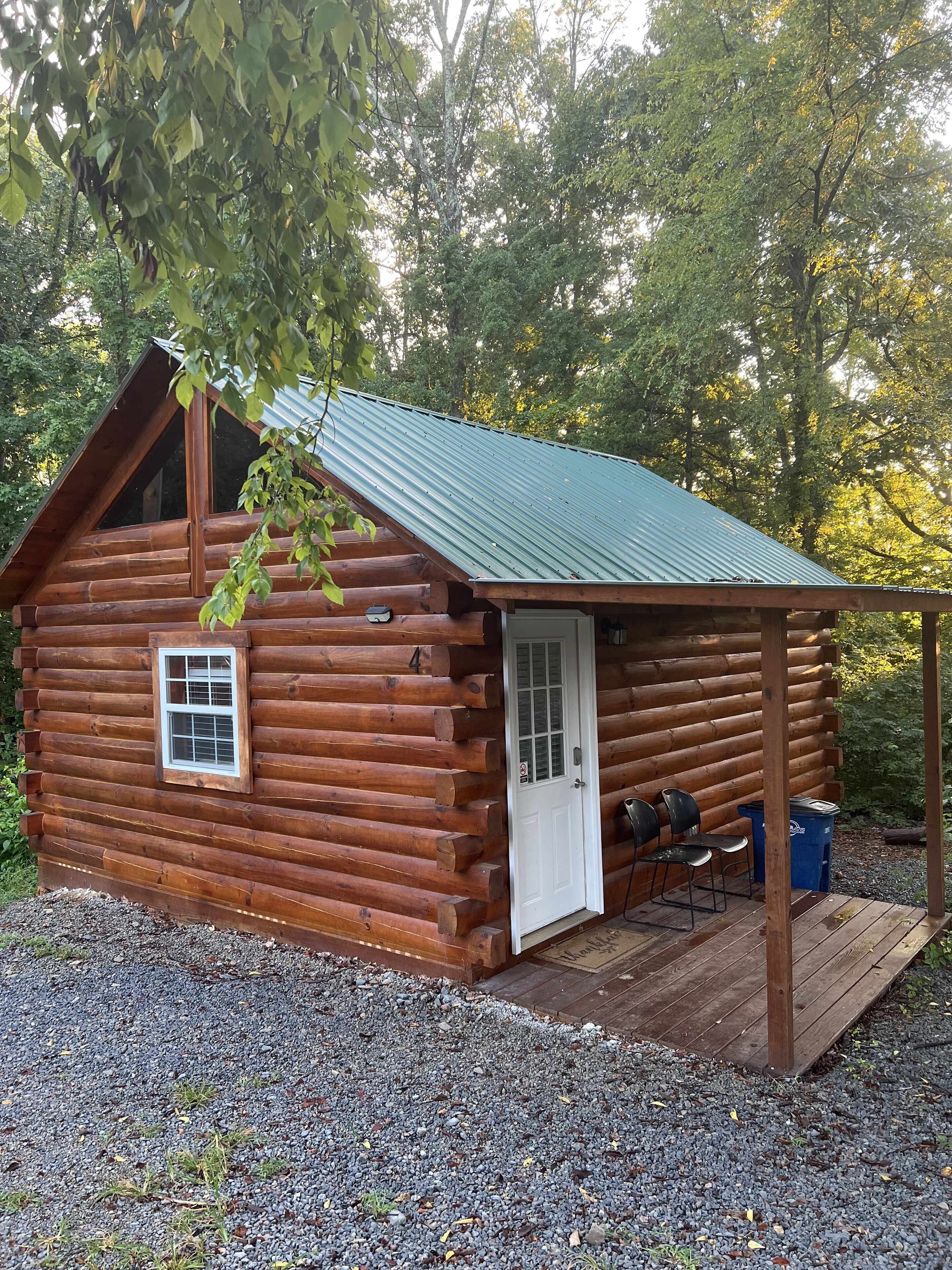 A log cabin with a metal roof, a front door, and a small porch area with two chairs and a trash can.