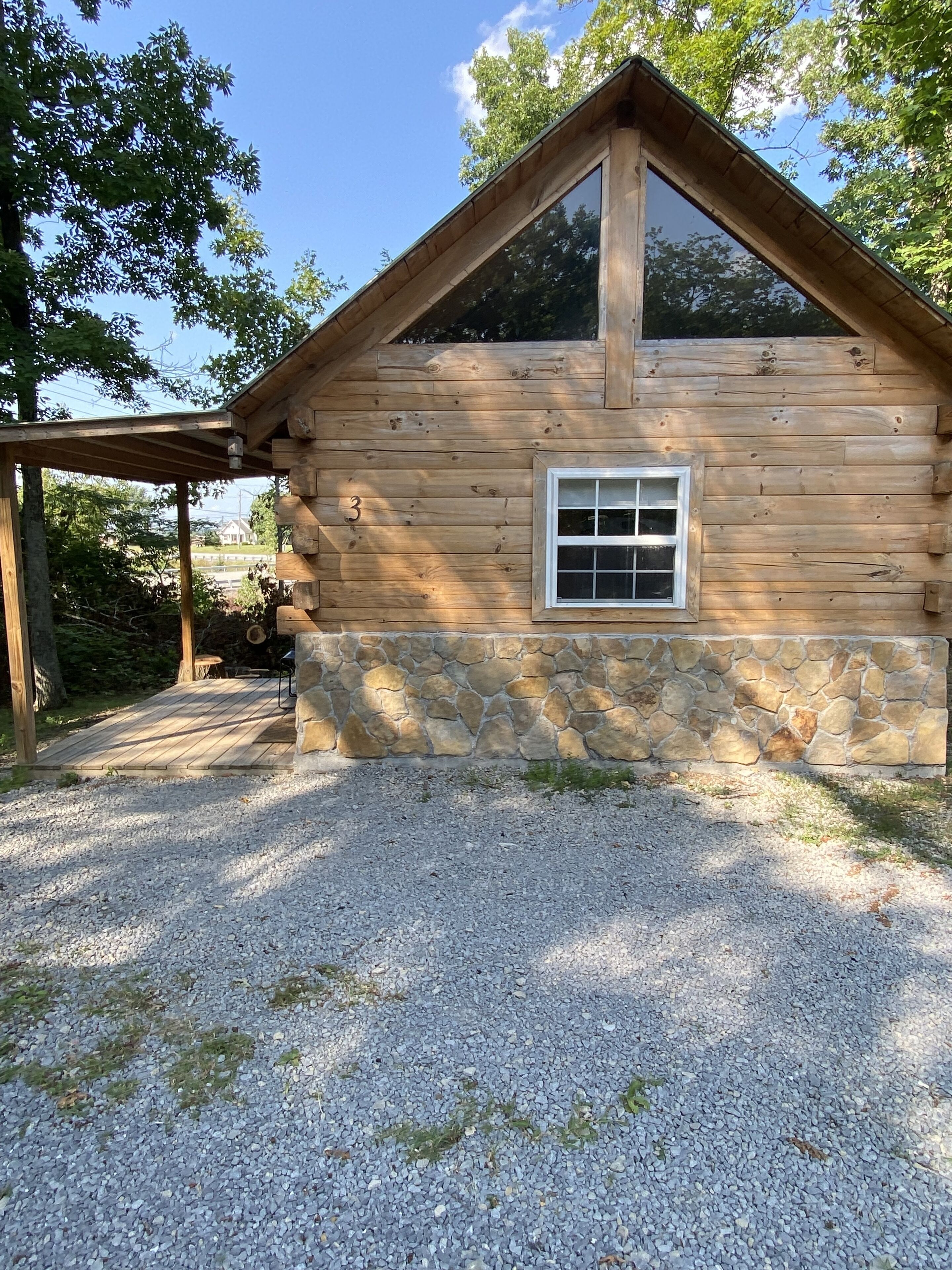 A log cabin with a stone foundation, a large window, and a gravel driveway.