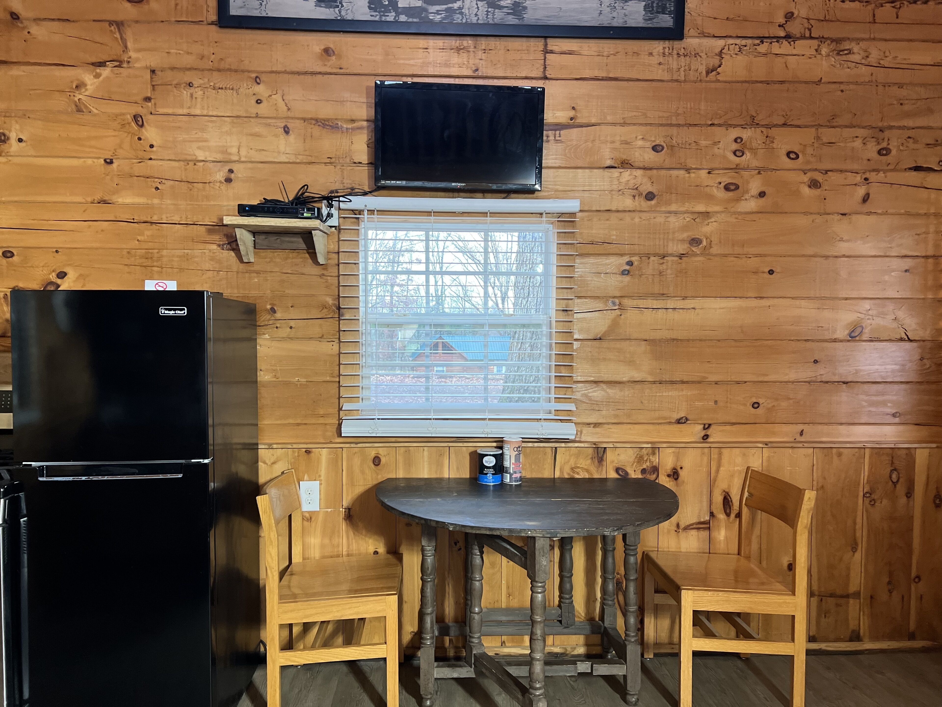 L’intérieur d’une cabane en bois, avec un réfrigérateur noir, une petite table avec deux chaises, une fenêtre avec des stores et un téléviseur fixé au mur.
