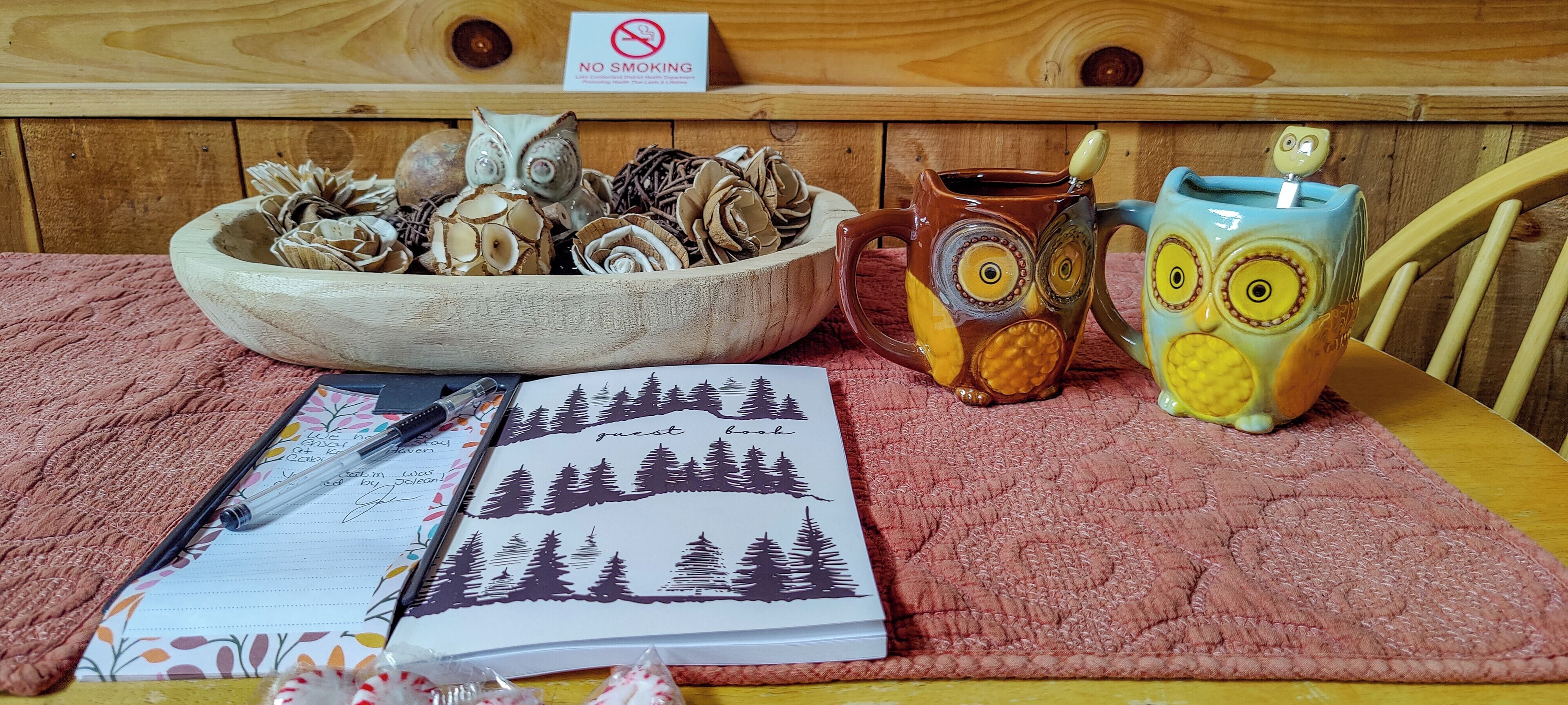 Une table en bois sur laquelle se trouvent un carnet, un bol rempli de petites décorations en forme de hibou et deux tasses également en forme de hibou.