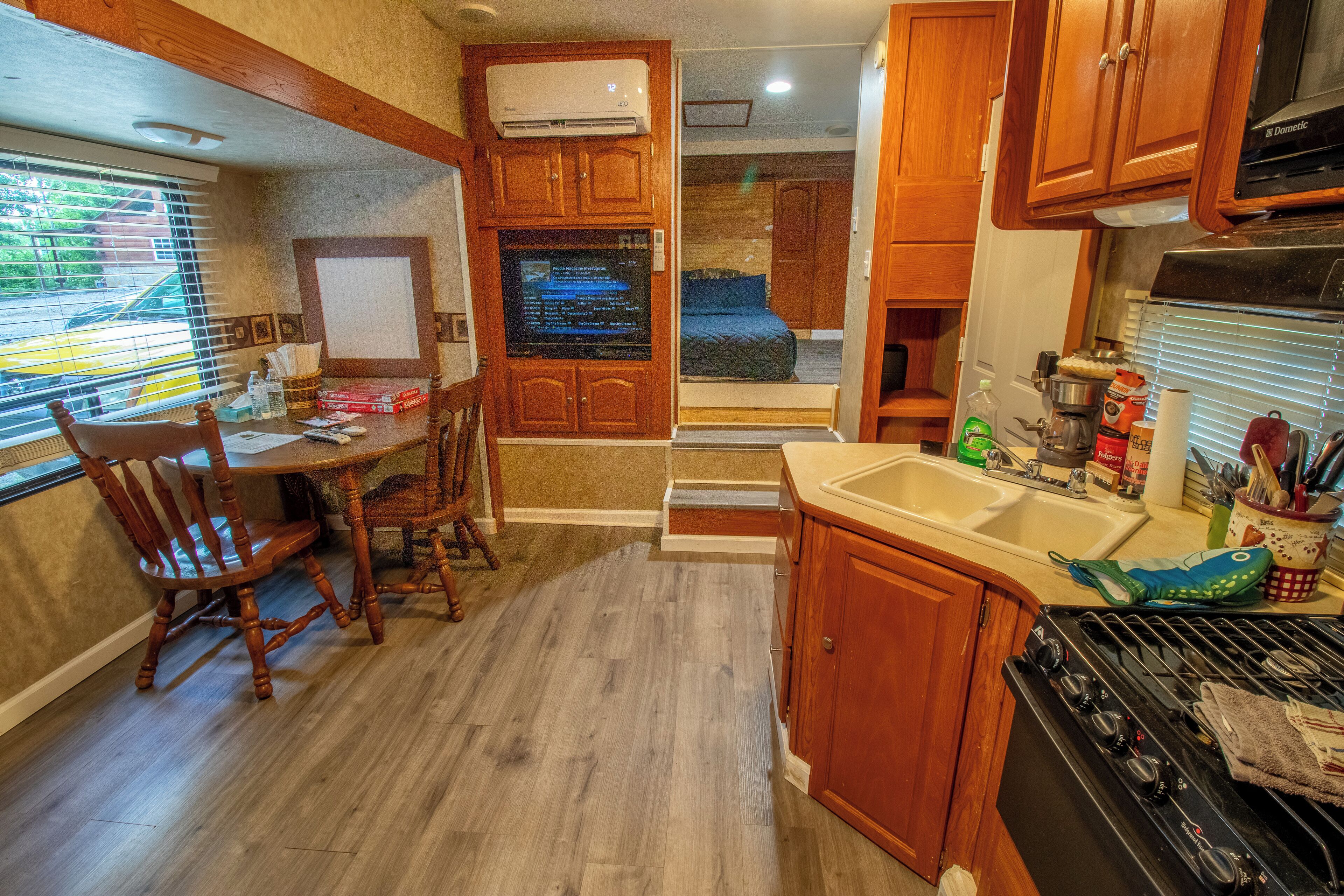 A compact kitchen with wooden cabinets, a dining table with chairs, a built-in oven, and a window with blinds.