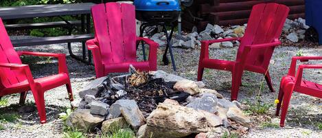 A campsite with a fire pit surrounded by red chairs, a blue grill, and a picnic table.