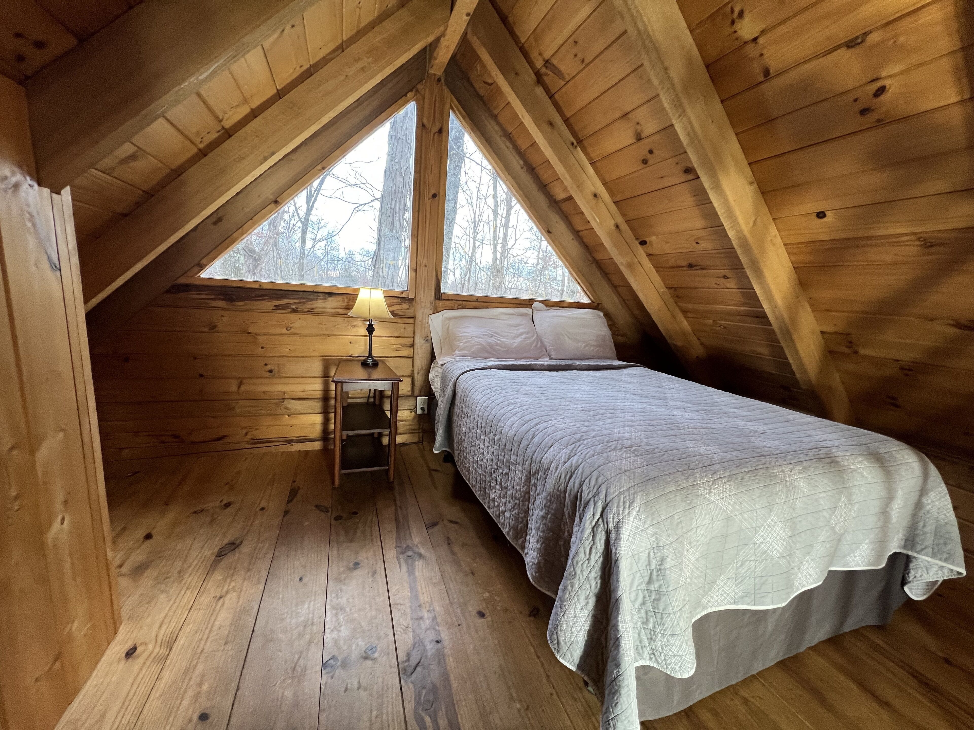 A cozy attic bedroom with a wooden sloped ceiling, a single bed with a quilted blanket, a nightstand with a lamp, and a window overlooking a forested area.