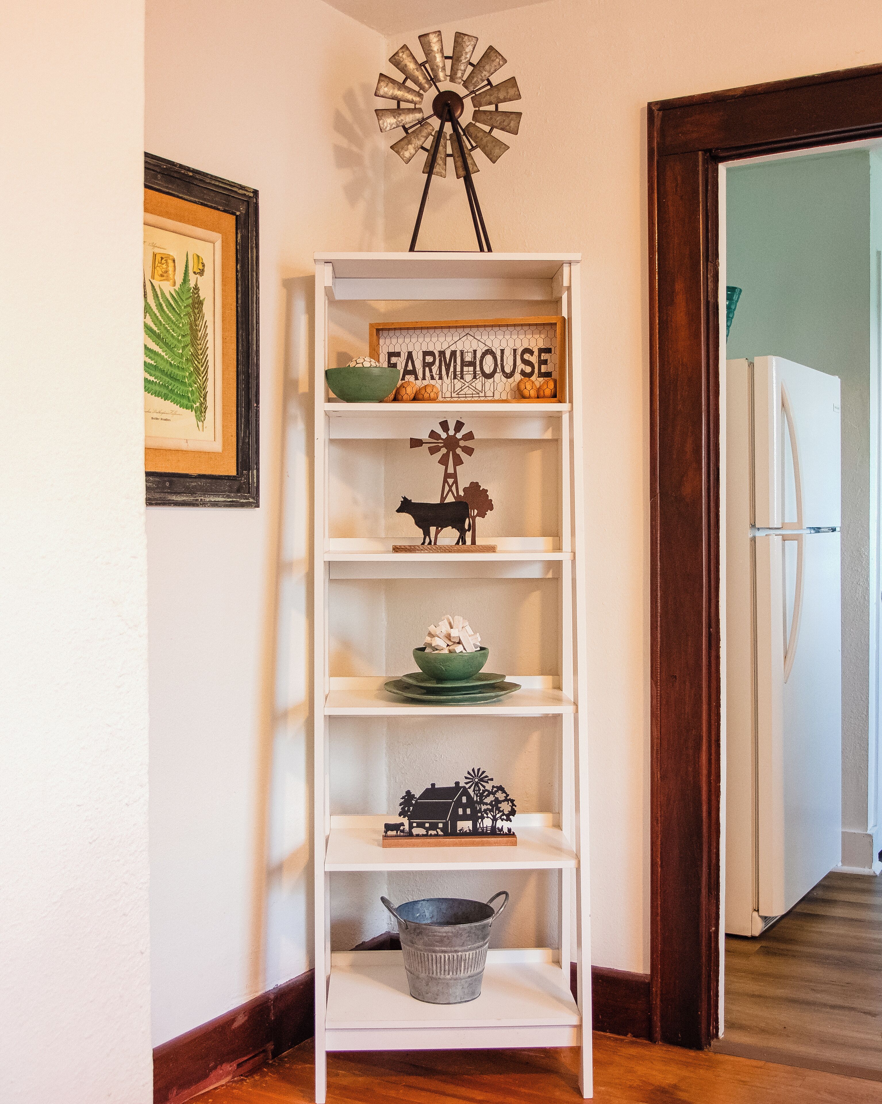 Una estantería blanca con objetos decorativos, incluyendo un molino, un cartel de granja y un cubo de metal.