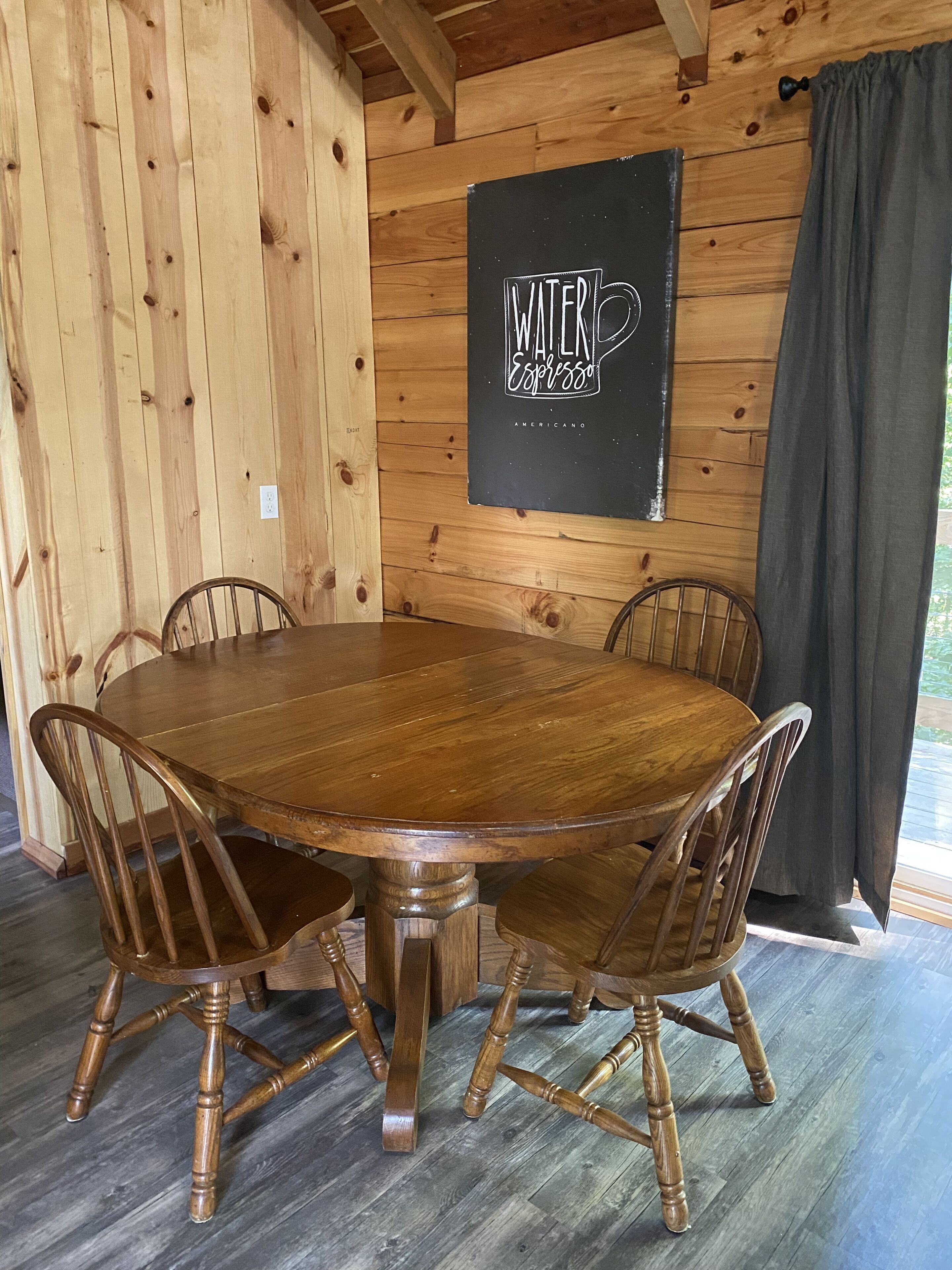 A wooden dining table with six chairs, a black sign on the wall, and a window with curtains.