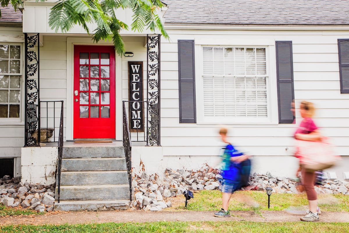 Uma casa com porta vermelha, um letreiro 'WELCOME' e degraus que levam à frente.