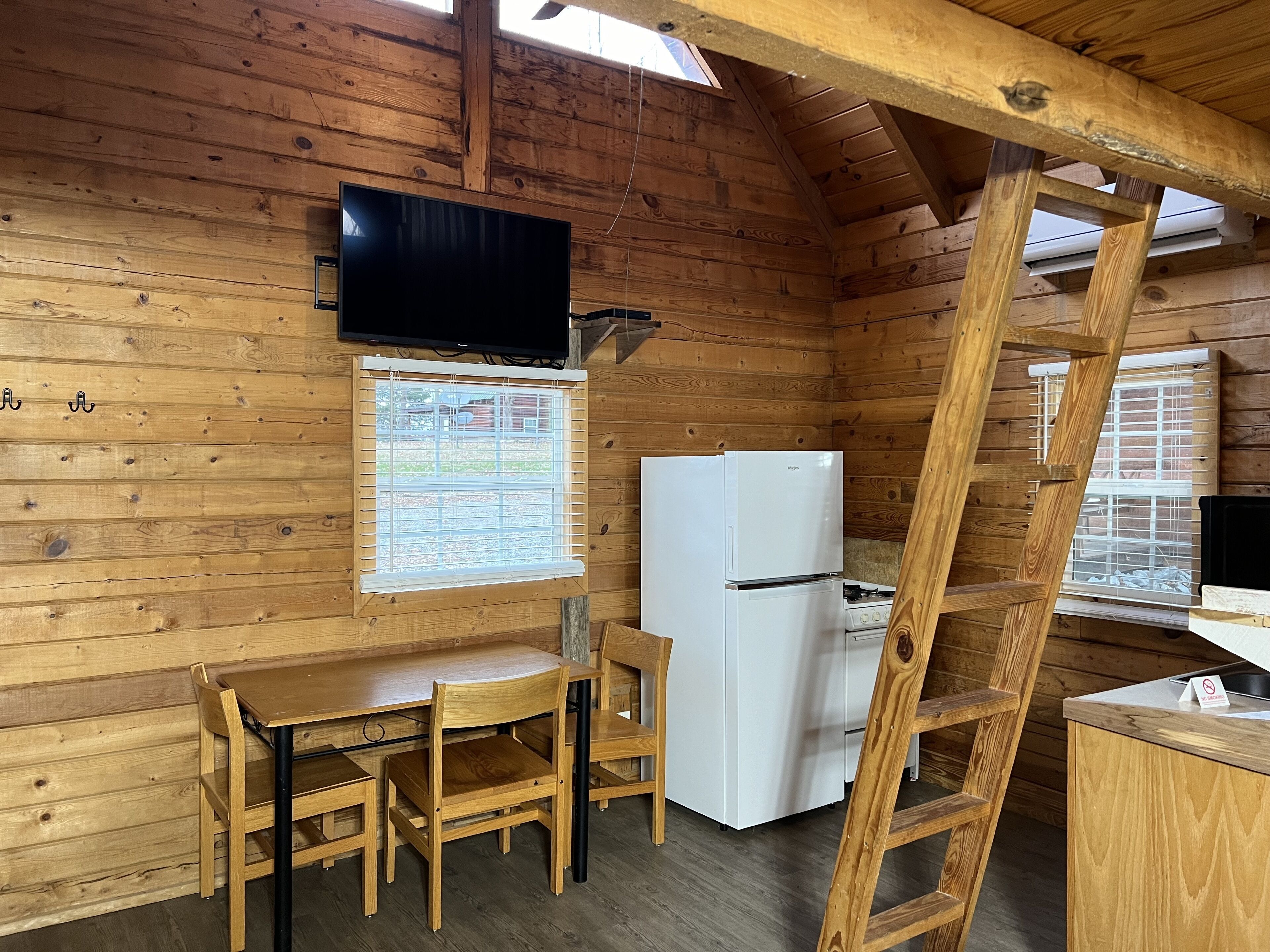 A wooden cabin interior with a dining table, chairs, a refrigerator, a stove, a TV mounted on the wall, and a window with blinds.