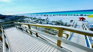 Ein Holzsteg führt zu einem Strand mit Sonnenschirmen und Menschen.