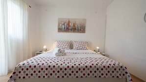A neatly made bed with a polka dot bedspread, a cityscape painting above it, and two bedside lamps. - Spacious apartment w. balcony, big garden & grill (Podglavica)