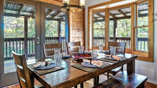 A wooden dining table set with chairs, placemats, and glassware, with large windows offering a view of the outdoors. - Amazing Wild Escape!-3Full Baths,Game,Hot Tub,Pets (Sevierville)