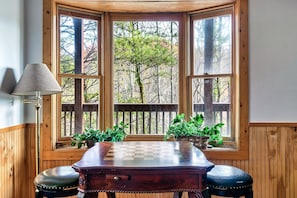 A wooden table with a chessboard, two stools, a lamp, and a view of trees through a large window with a wooden frame.