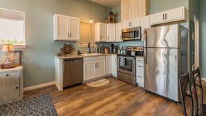 Une cuisine avec des meubles blancs, des appareils en acier inoxydable, un plancher en bois et une fenêtre avec des stores.