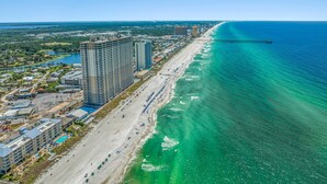An aerial view of a coastal area with a long sandy beach, a pier extending into the ocean, and a large hotel complex with multiple buildings and a swimming pool. - 𝒫𝒞𝐵 𝒫𝒶𝓇𝒶𝒹𝒾𝓈𝑒 4 BR, 3BA (Panama City Beach)