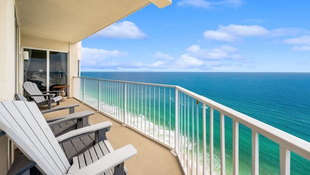 A balcony with Adirondack chairs, a glass door leading to a room, and a view of the ocean. - 𝒫𝒞𝐵 𝒫𝒶𝓇𝒶𝒹𝒾𝓈𝑒 4 BR, 3BA (Panama City Beach)
