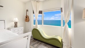 A bedroom with a bed, a nightstand, a lamp, a guitar, a window with curtains, and a view of the ocean. - 𝒫𝒞𝐵 𝒫𝒶𝓇𝒶𝒹𝒾𝓈𝑒 4 BR, 3BA (Panama City Beach)