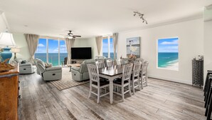 A spacious living room with a large dining table, comfortable seating, a flat-screen TV, and large windows offering ocean views. - 𝒫𝒞𝐵 𝒫𝒶𝓇𝒶𝒹𝒾𝓈𝑒 4 BR, 3BA (Panama City Beach)