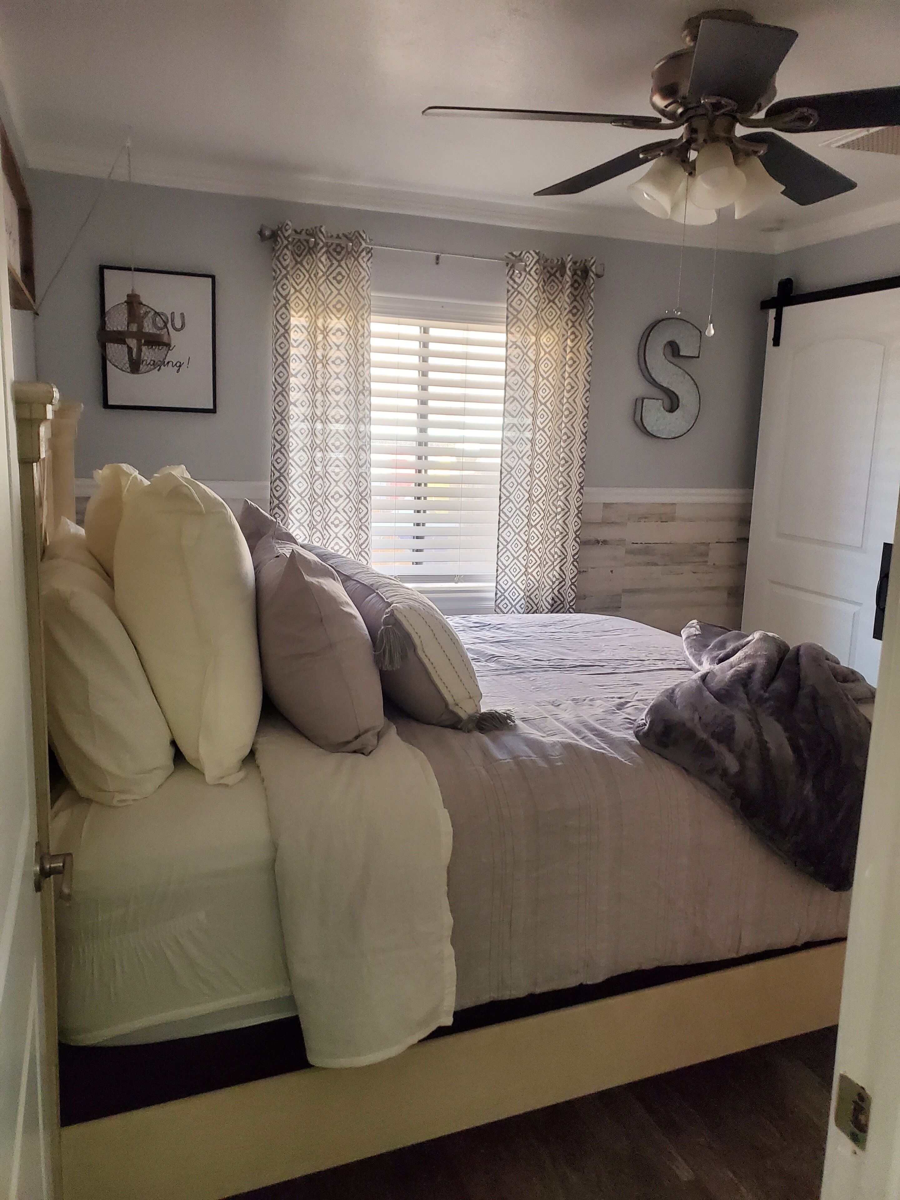 A bedroom with a bed, pillows, a ceiling fan, a window with blinds, and a wall with a letter decoration.