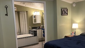 Ein Schlafzimmer mit Bett, Nachttisch, Lampe, Deckenventilator und einem durchgegangenen Türspalte sichtbaren Badezimmer.