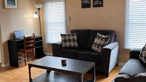 Ein Wohnzimmer mit Deckenventilator, einer Sofagarnitur, einem Couchtisch, einem Schreibtisch mit Laptop und zwei an der Wand befestigten Bilderrahmen.