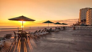 Una terraza frente a la playa con tumbonas y sombrillas, con vistas al océano al atardecer.
